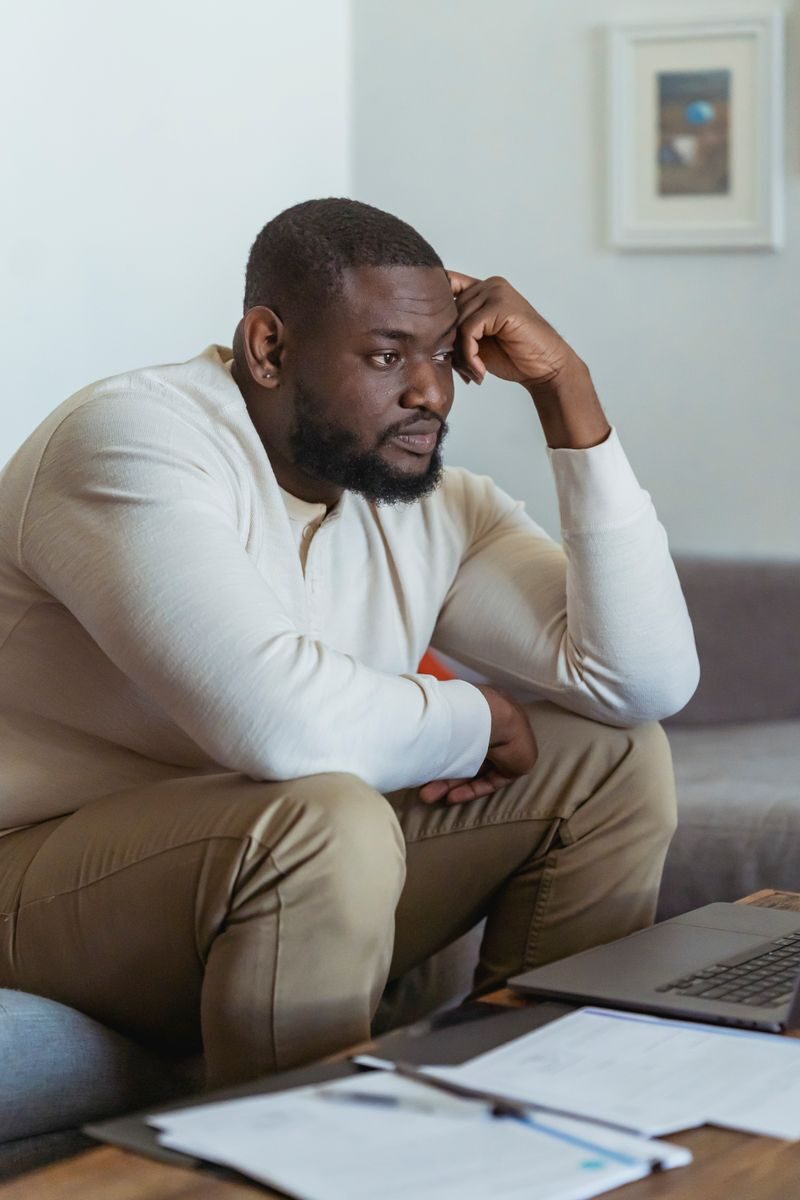 Depressed African American sitting on soft couch near table with papers and laptop and pondering